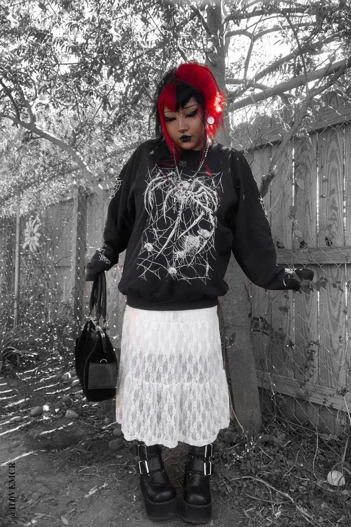 Person with red hair wearing a black hoodie with a white design, white skirt, and black shoes in a forest setting.