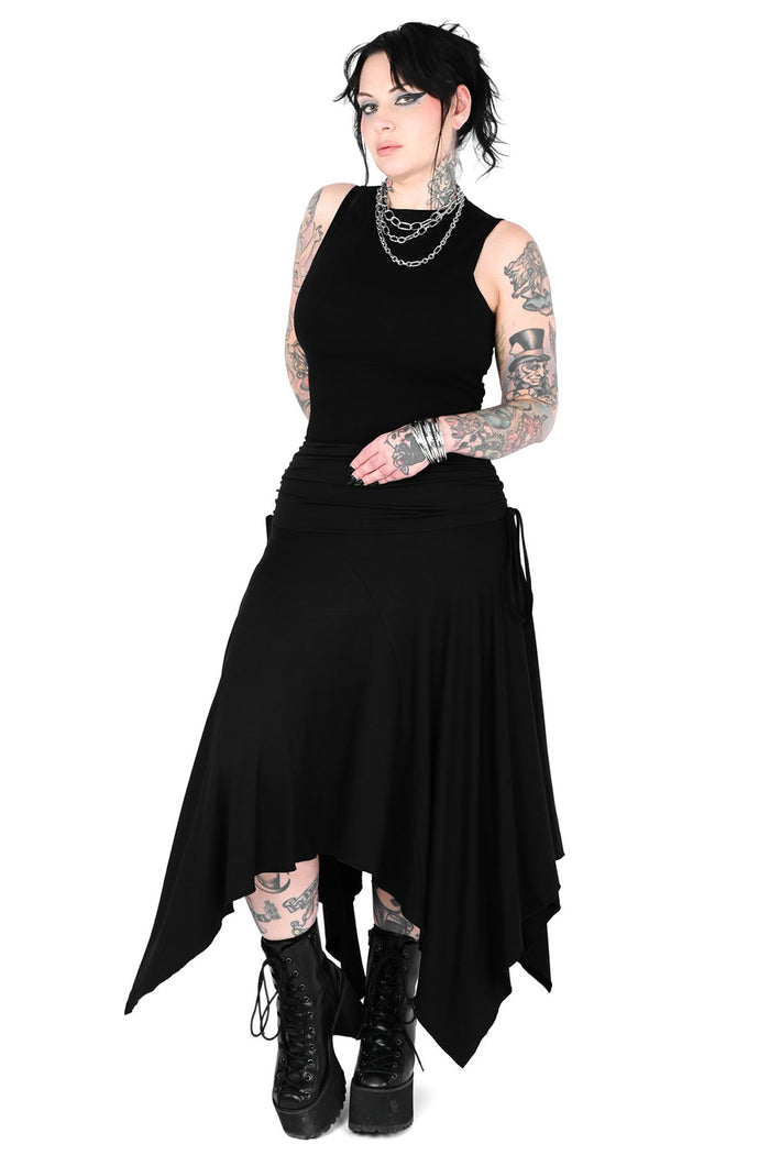 A model with tattoos is wearing a black sleeveless high-low skirt with an asymmetrical hem, layered with silver chain necklaces and black platform combat boots, set against a plain white background.