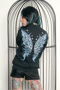 A person with black and teal hair stands in front of a black metal birdcage, wearing a black sweatshirt with a large light blue and white wing graphic on the back, paired with black shorts.
