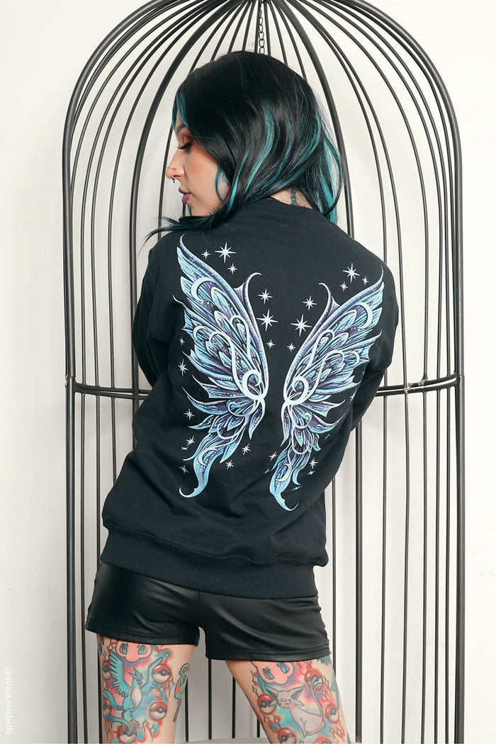 A person with black and teal hair stands in front of a black metal birdcage, wearing a black sweatshirt with a large light blue and white wing graphic on the back, paired with black shorts.