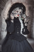 A gothic-style model wearing a black layered ruffle top, a large black lace hat with a veil, and a cross necklace, posed against a dark, atmospheric stone background.