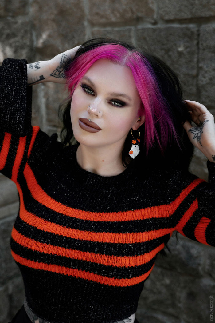 Person with pink hair wearing a black and orange striped sweater against a stone wall with ghost earrings